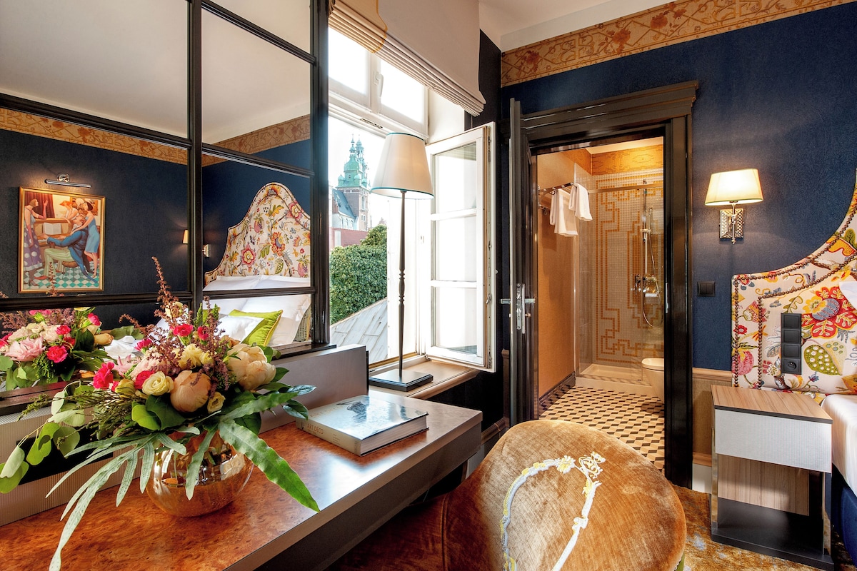 Quarto de hotel com um espelho grande, uma escrivaninha de madeira com um vaso de flores, uma cama com cabeceira estampada e vista para a cidade pela janela.