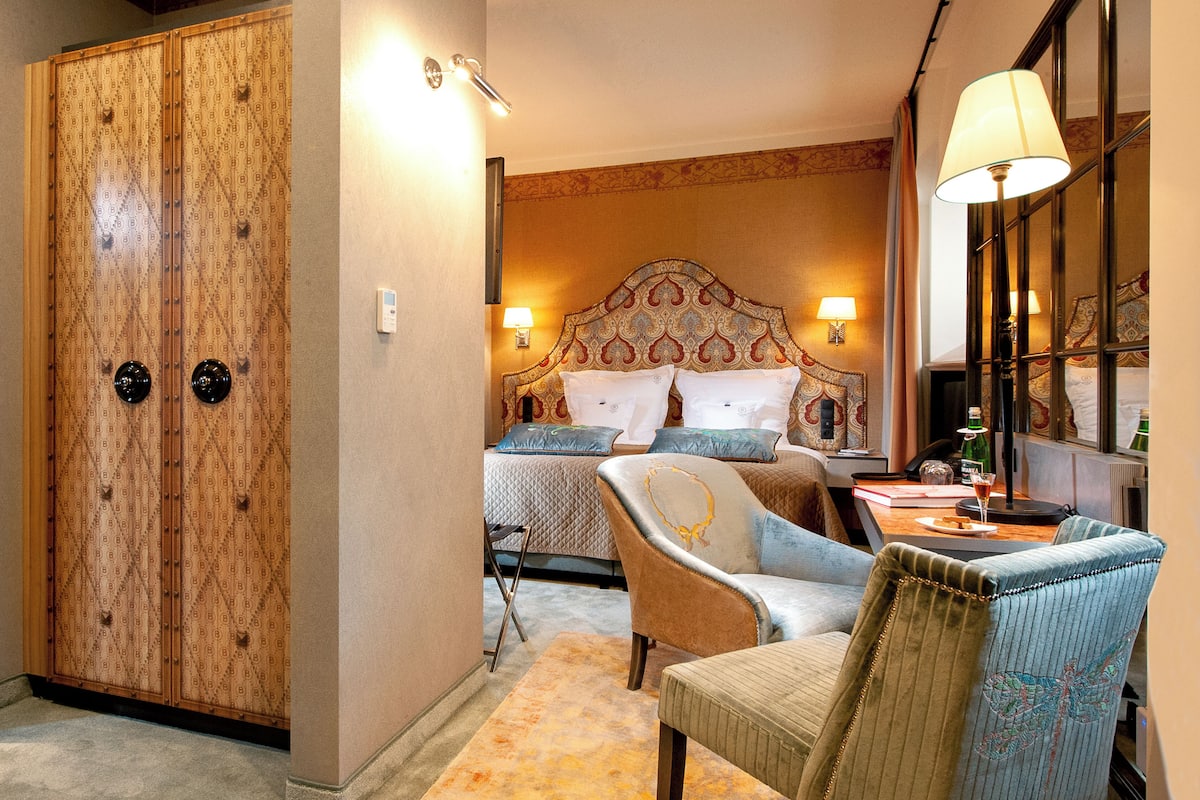 A hotel room with a large wardrobe, a bed with patterned bedding, two chairs, a table with a bottle and glasses, and a lamp.