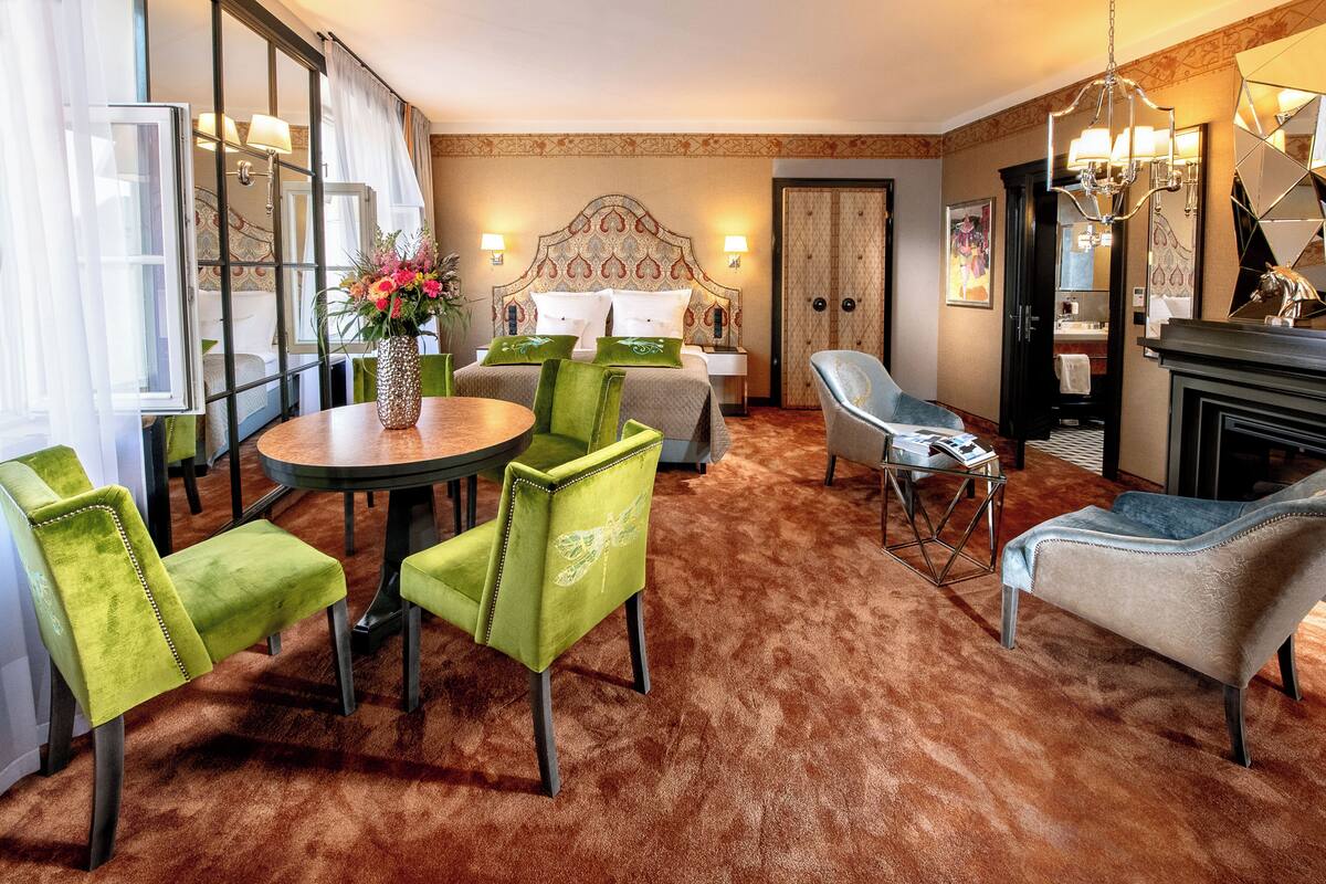 A hotel room with a large bed, a sitting area with a table and chairs, a bathroom, and a view through a large window.