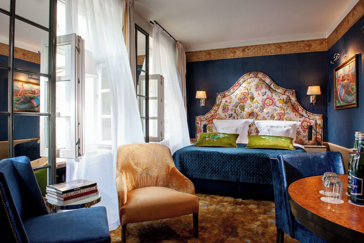 A hotel room with a large bed, a chair, a small table, and a bookshelf.