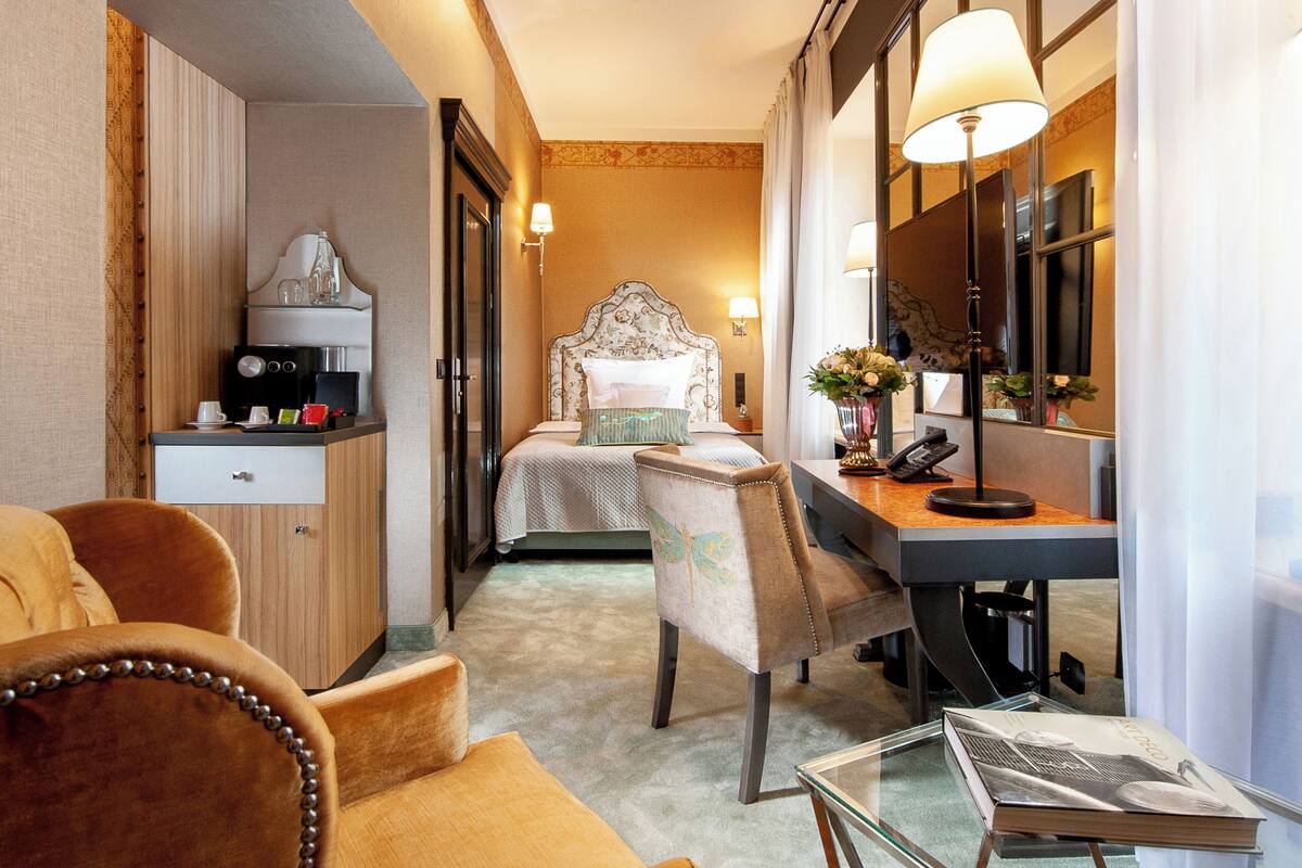 A compact hotel room with a bed, a desk, a chair, a coffee machine, and a small table with a magazine.