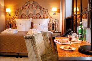Une chambre d’hôtel avec un grand lit, deux lampes de chevet, une table de chevet avec un téléphone, une table avec un verre de vin, une bouteille de vodka Smirnoff et une chaise.
