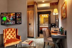 Une chambre d’hôtel dotée d’un grand téléviseur à écran plat, d’un fauteuil capitonné, d’une petite table avec un livre, d’un bureau avec une lampe et d’une salle de bain visible à travers une porte ouverte.