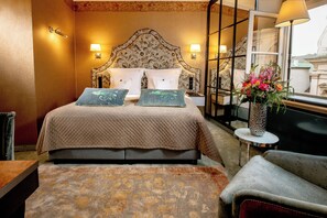 A hotel room with a large bed, two bedside lamps, a vase with flowers, a chair, and a view of a building through a window. - Balthazar Design Hotel (Kraków)