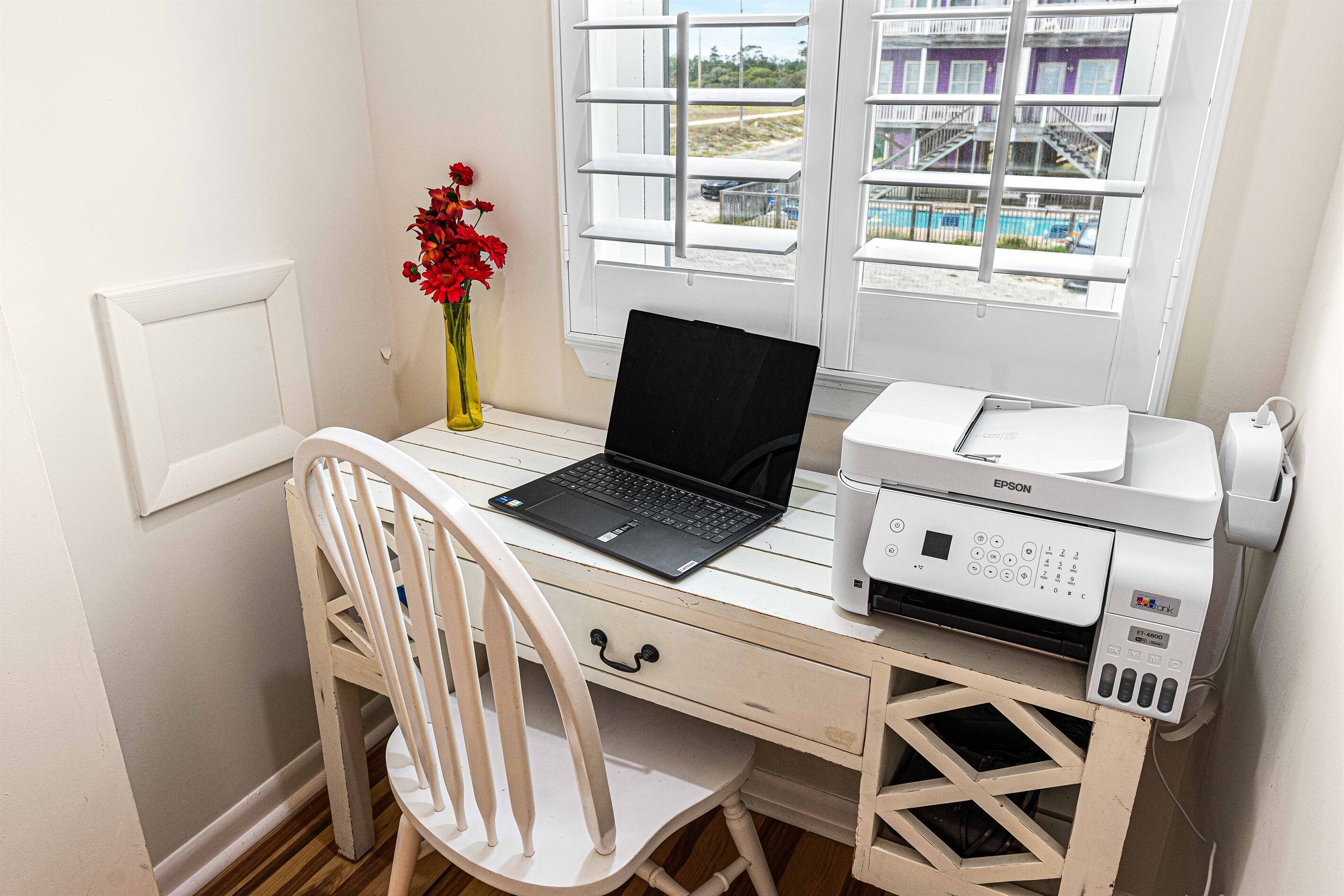 A white desk with a laptop, a printer, and a vase with red flowers.
