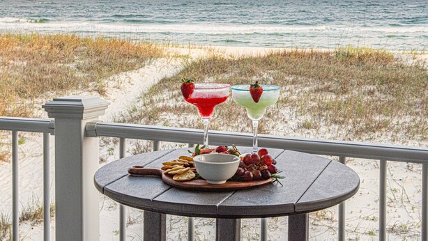 Uma mesa com uma tigela de uvas, um prato de biscoitos e dois coquetéis numa varanda com vista para a praia e o oceano ao pôr do sol.