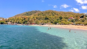 Ein Strand mit klarem türkisblauem Wasser, badende Menschen und ein bewaldeter Hügel im Hintergrund.