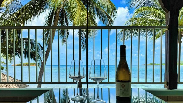 Ein Balkon mit Glastisch, zwei Weingläsern und einer Weinflasche, mit Blick auf einen Strand mit Palmen und das Meer.