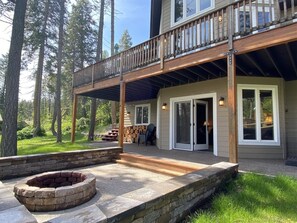 A two-story house with a covered patio, stone fire pit, and a deck with a railing. - Gt254 Goat Trail Lookout (Whitefish)