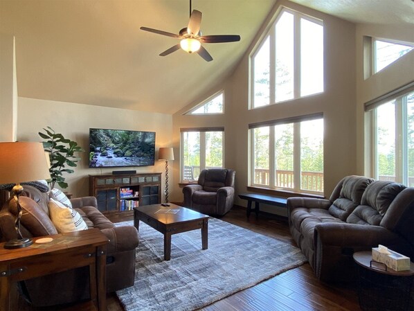 A spacious living room with a ceiling fan, large windows, a flat-screen TV, leather sofas, a coffee table, and a bookshelf. - Gt254 Goat Trail Lookout (Whitefish)