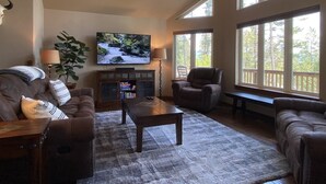 A spacious living room with a large flat-screen TV mounted on the wall, a comfortable sofa set, a wooden coffee table, and a large window offering a view of trees. - Gt254 Goat Trail Lookout (Whitefish)