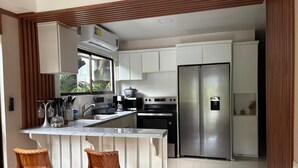 A modern kitchen with white cabinets, stainless steel appliances, a built-in oven, and a refrigerator. There are two wooden bar stools and a window with a view of greenery. - Arenal Poshpacker Boutique Urban Tropical Hotel (La Fortuna)