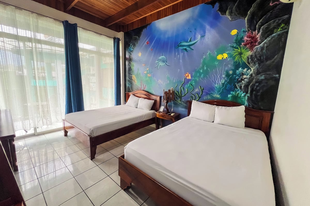 A hotel room with two beds, a mural of an underwater scene, and a view through a window with curtains.