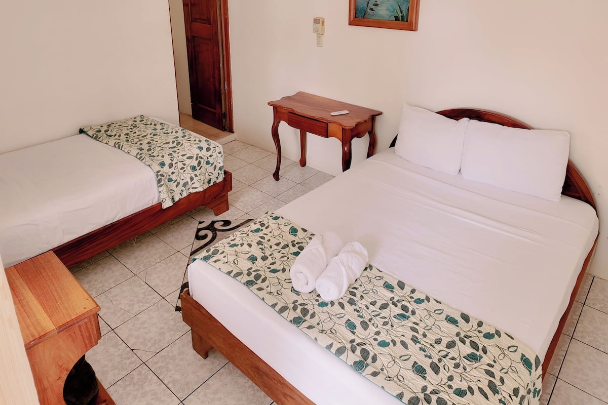 A hotel room with two single beds, a wooden nightstand, a desk, a painting, and a ceiling fan.