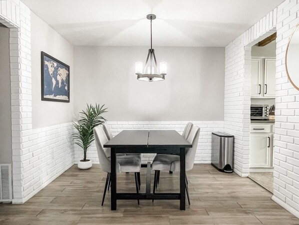 Uma área de refeições com mesa preta, cadeiras brancas, uma planta em vaso e um lustre.