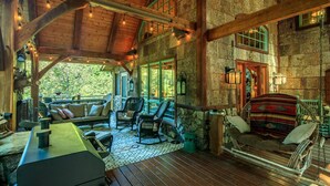 Interior rústico de una cabaña con terraza de madera, mobiliario, paredes de piedra y un ventanal con vistas a la vegetación.