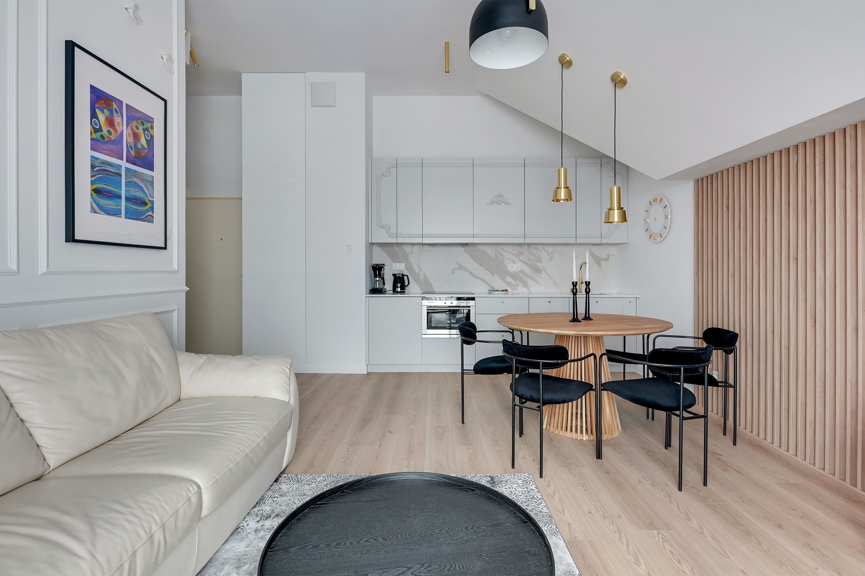 Un soggiorno moderno con divano, un'area pranzo con tavolo rotondo e sedie, e una cucina con mobili bianchi e parete in legno.