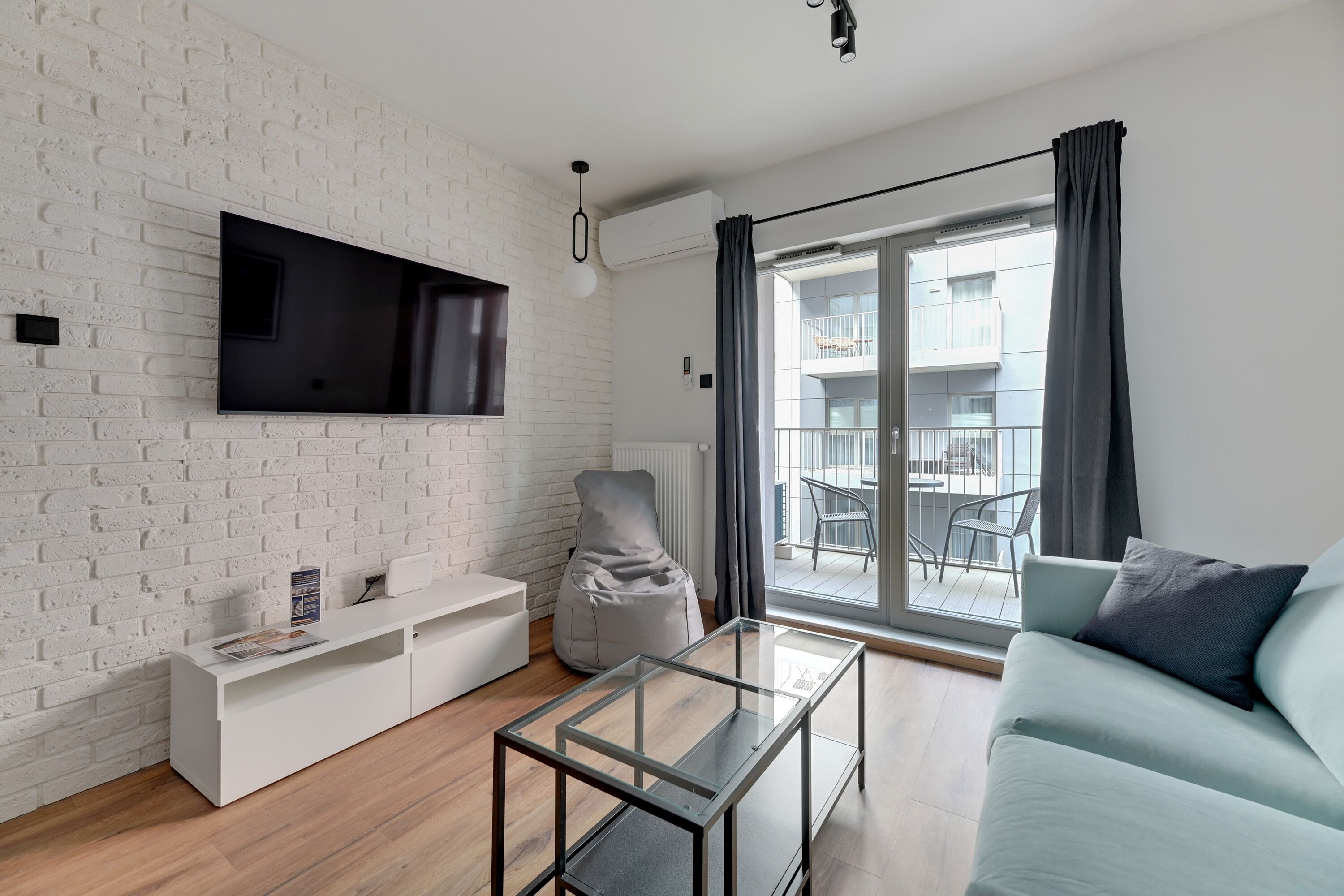 Un soggiorno moderno con parete in mattoni bianchi, TV a schermo piatto, tavolino in vetro, divano azzurro e balcone con vista sulla città.