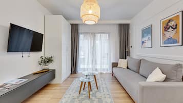 Una sala de estar moderna con un sofá gris, una mesa de centro de vidrio, un televisor montado en la pared y un gran ventanal con cortinas translúcidas.
