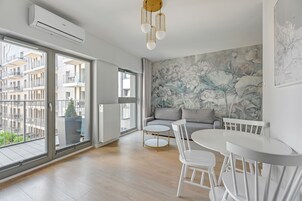 Una sala de estar moderna con un ventanal grande, un sofá gris, una mesa de comedor blanca con sillas, y un aire acondicionado instalado en la pared.