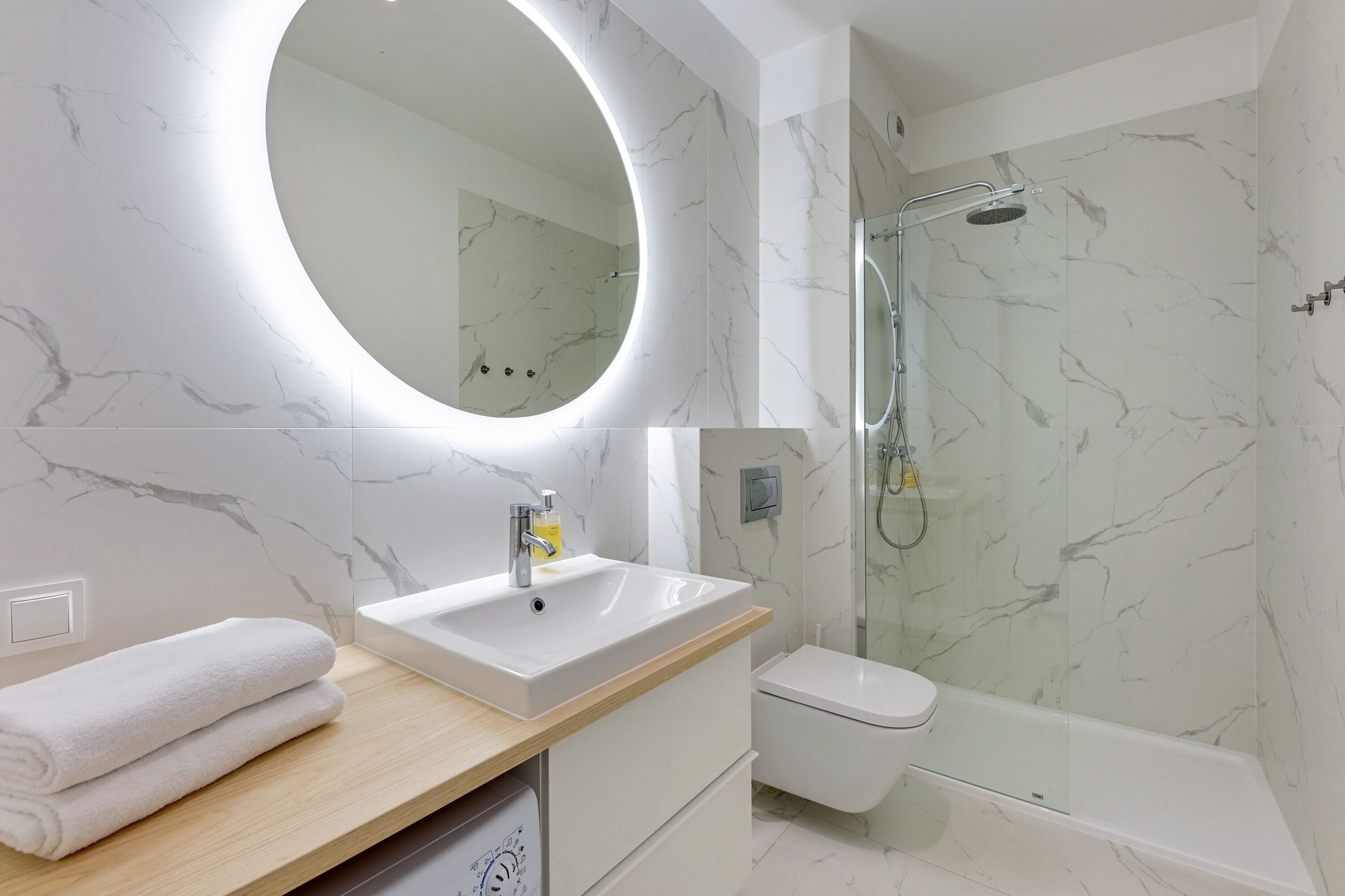 A modern bathroom with a marble vanity, a round mirror, a toilet, and a glass-enclosed shower.