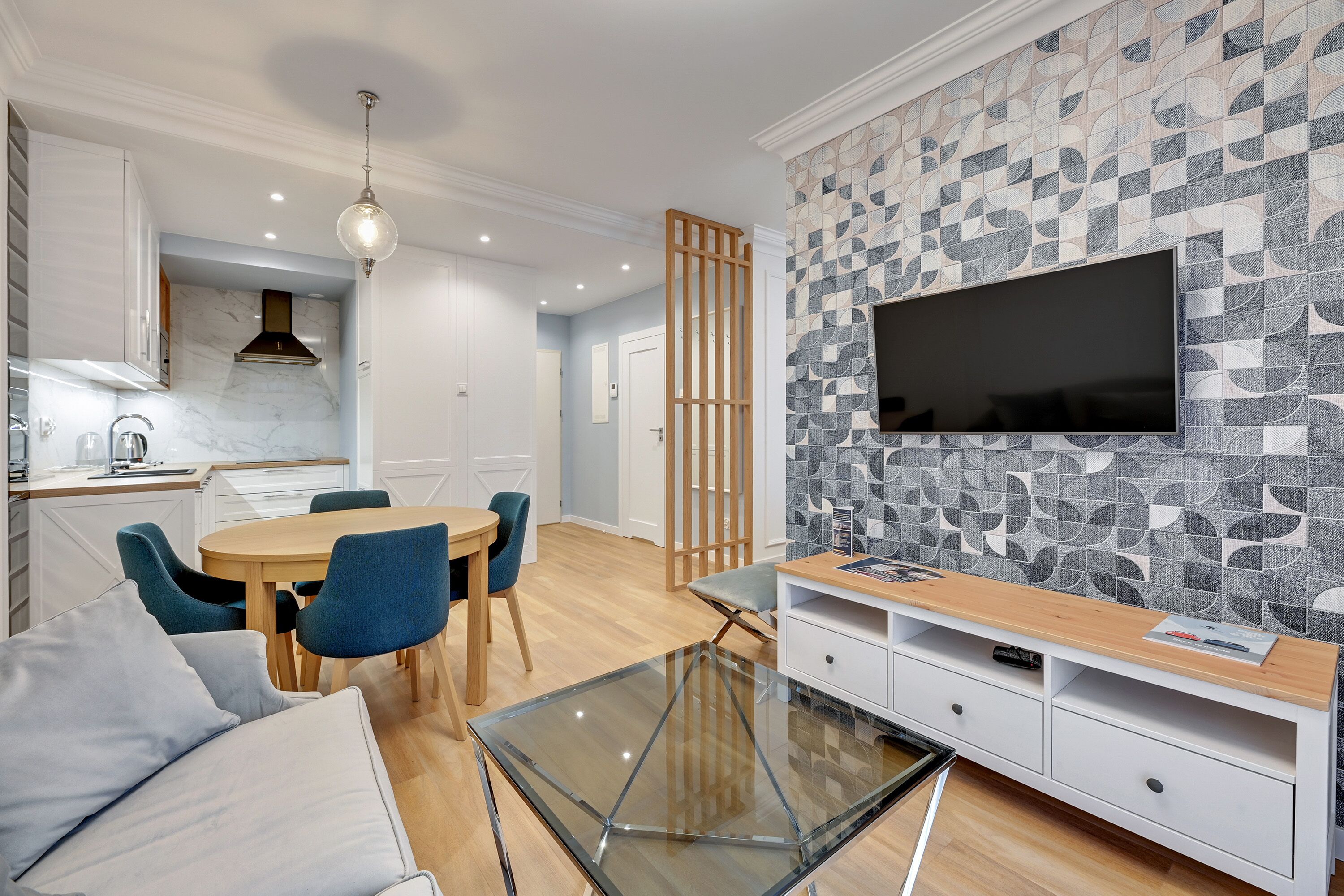 A modern living room with a glass coffee table, a sofa, a TV mounted on the wall, and a dining area with chairs and a table.