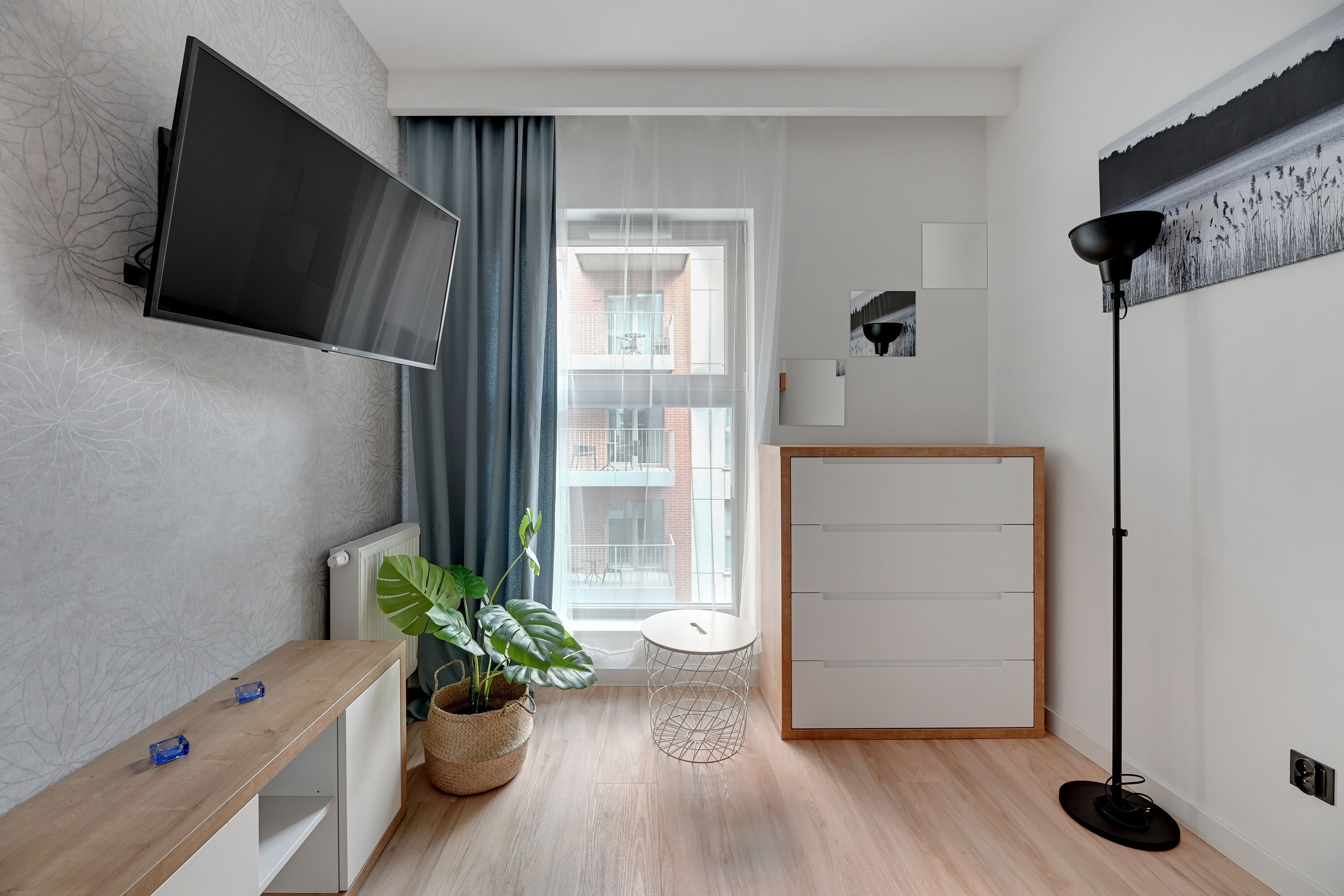 Habitación moderna y bien iluminada con un televisor grande, una pequeña mesa redonda, una planta en maceta, una lámpara de pie y vistas al balcón exterior.