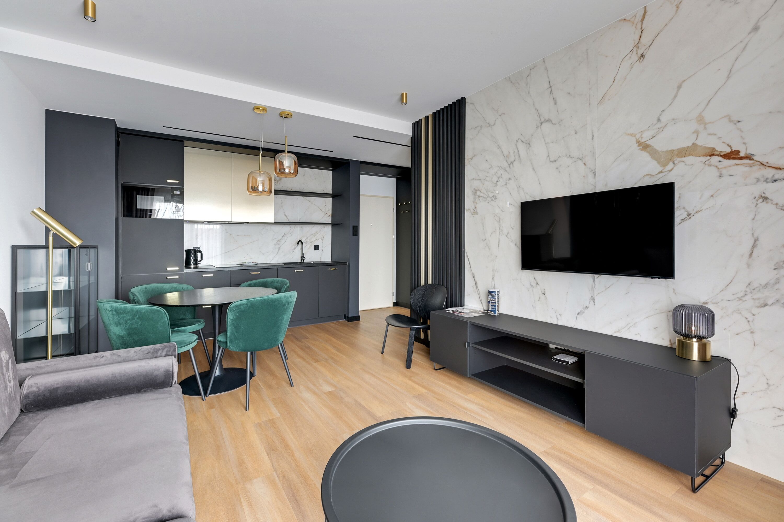 A modern living room with a sofa, dining table, and chairs, a kitchen area with cabinets and a TV mounted on the wall.