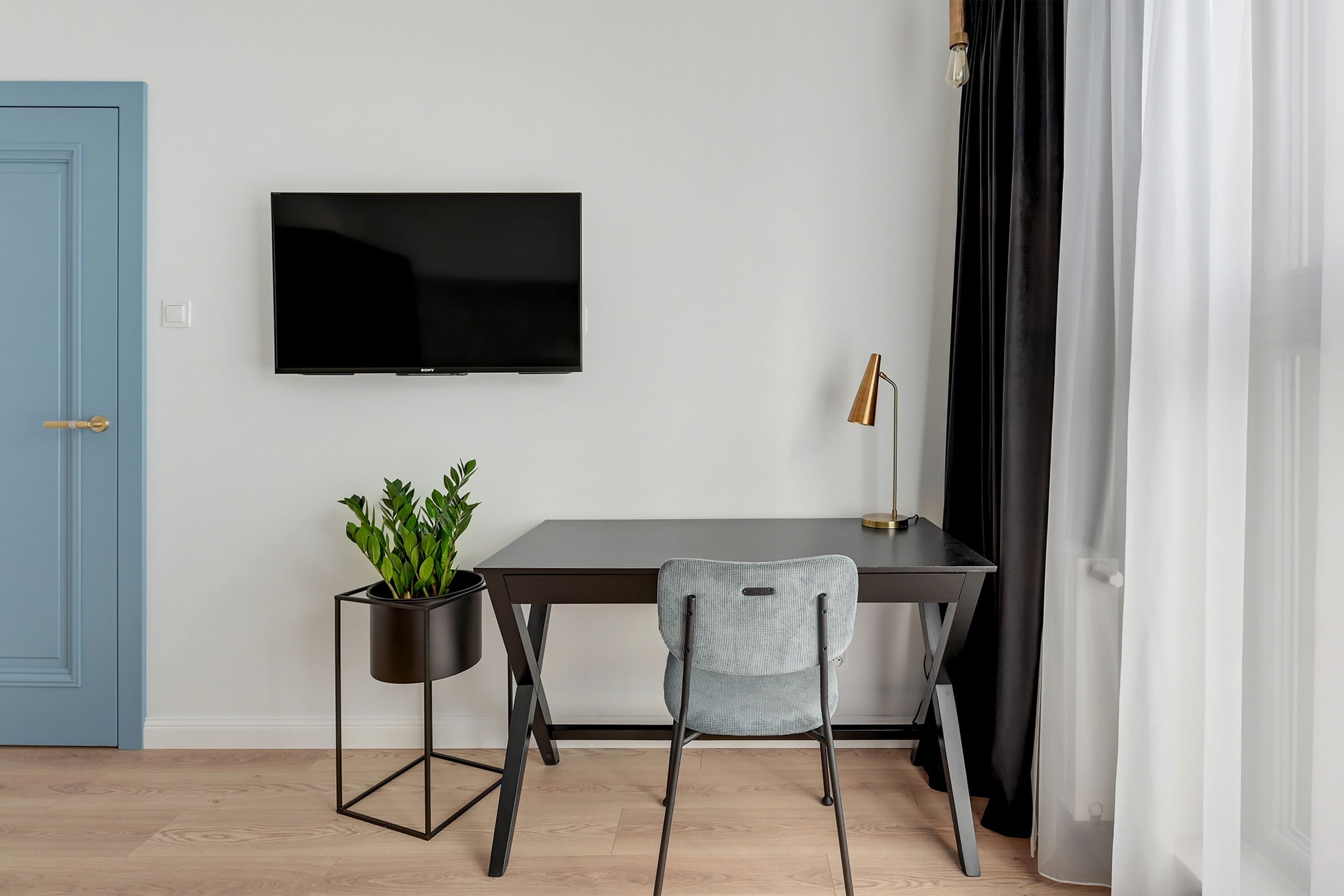 Et moderne værelse med et mørkt træskrivebord, et sort tv ophængt på væggen, en potteplante, en grå stol og en sort lampe.