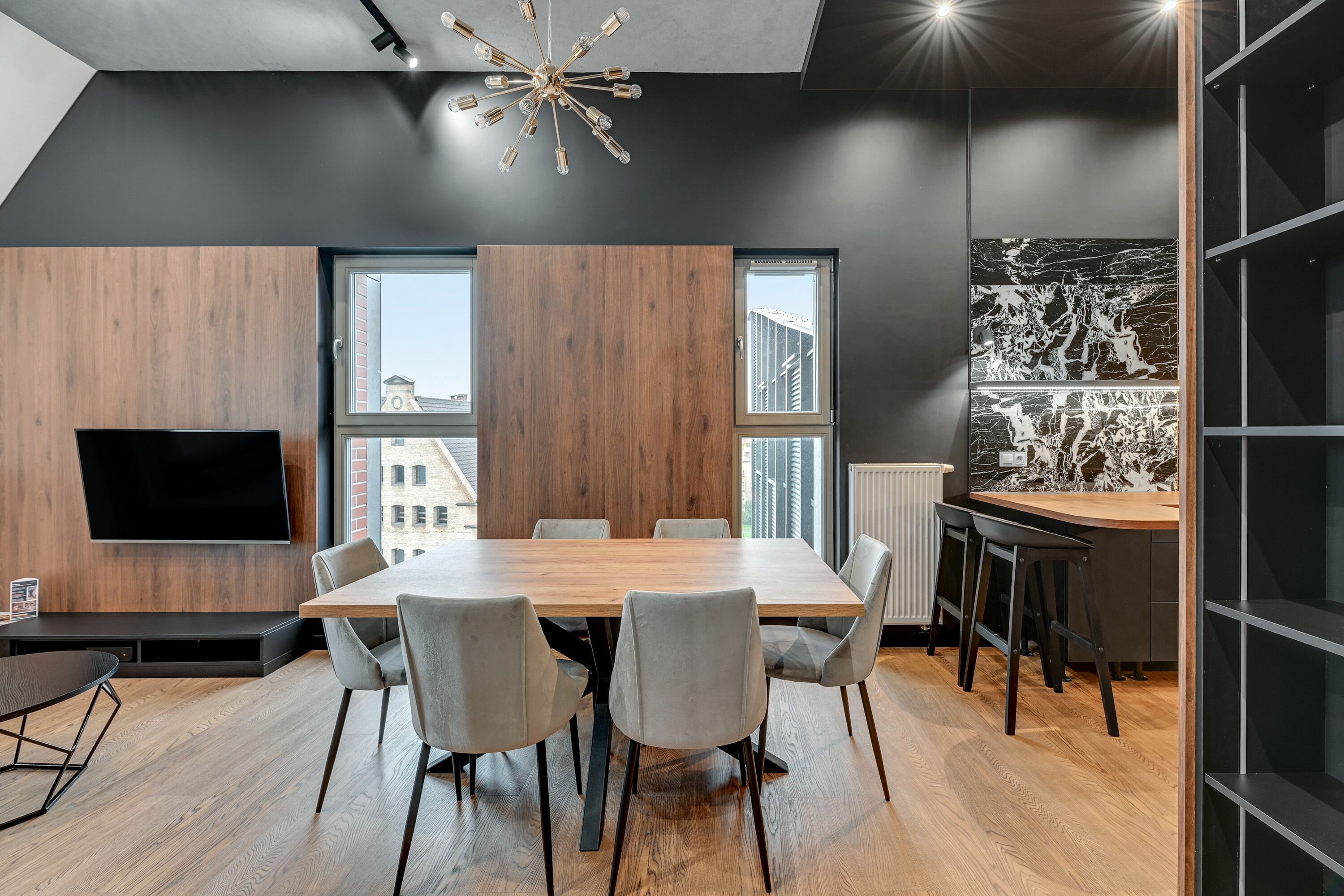 A modern conference room with a large wooden table, grey chairs, a flat-screen TV, and large windows offering city views.