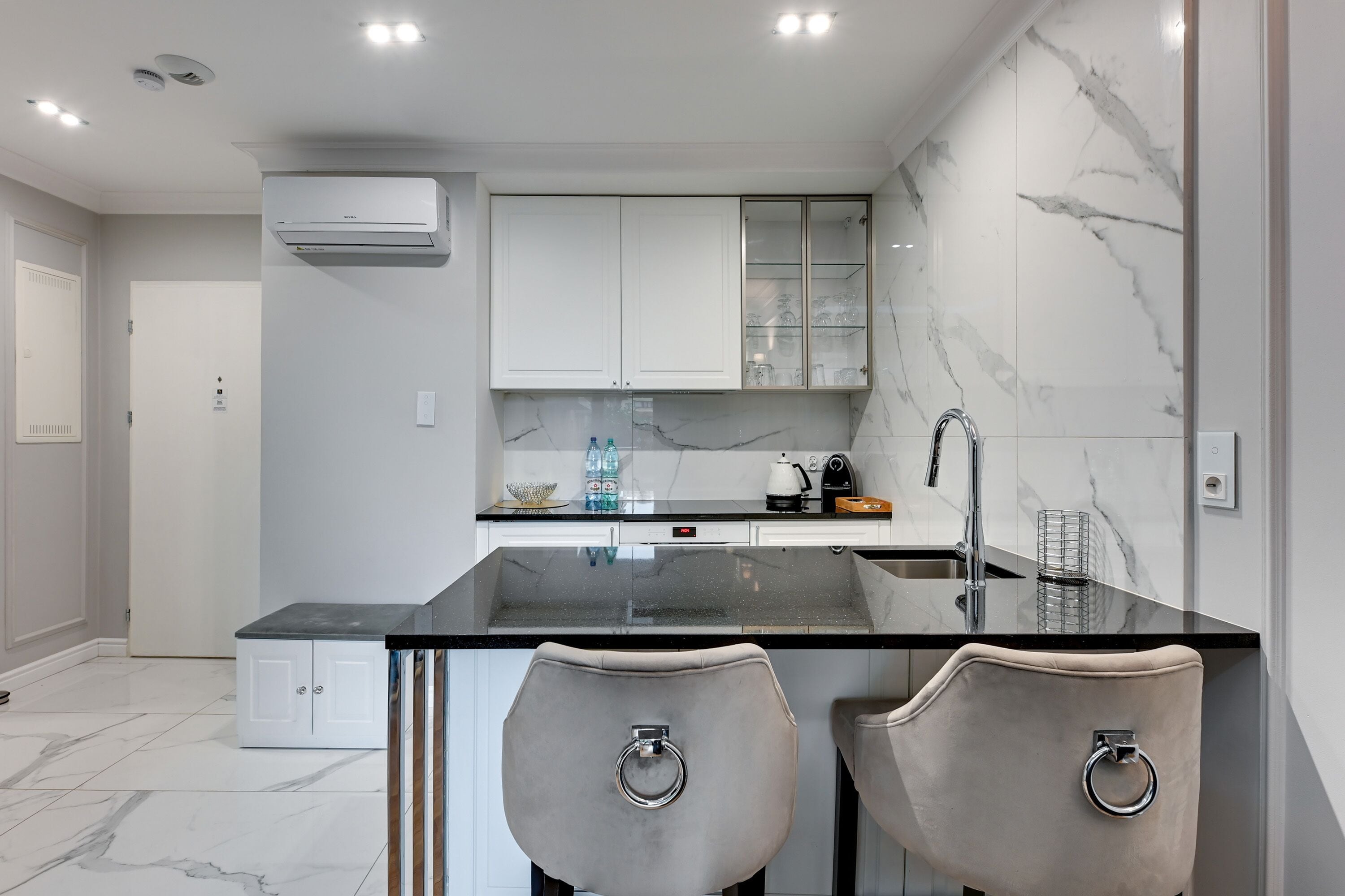 A modern kitchen with a marble backsplash, white cabinets, and a black countertop. There are two bar stools with metal rings.