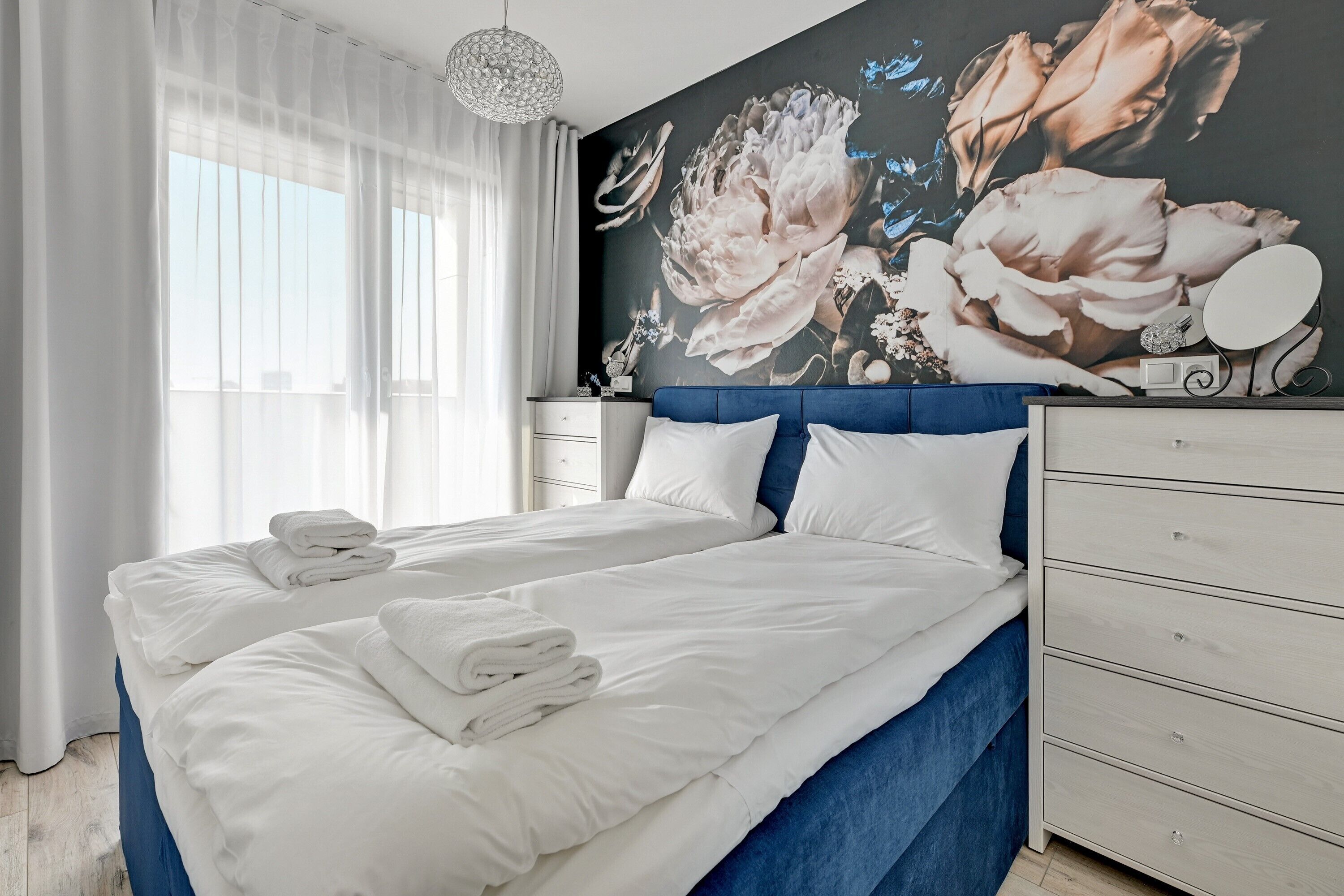 Ein ordentlich bezogenes Bett mit weißen Leinen und Handtüchern, ein blauer Kopfteil und eine Wand mit floralem Wandbild.