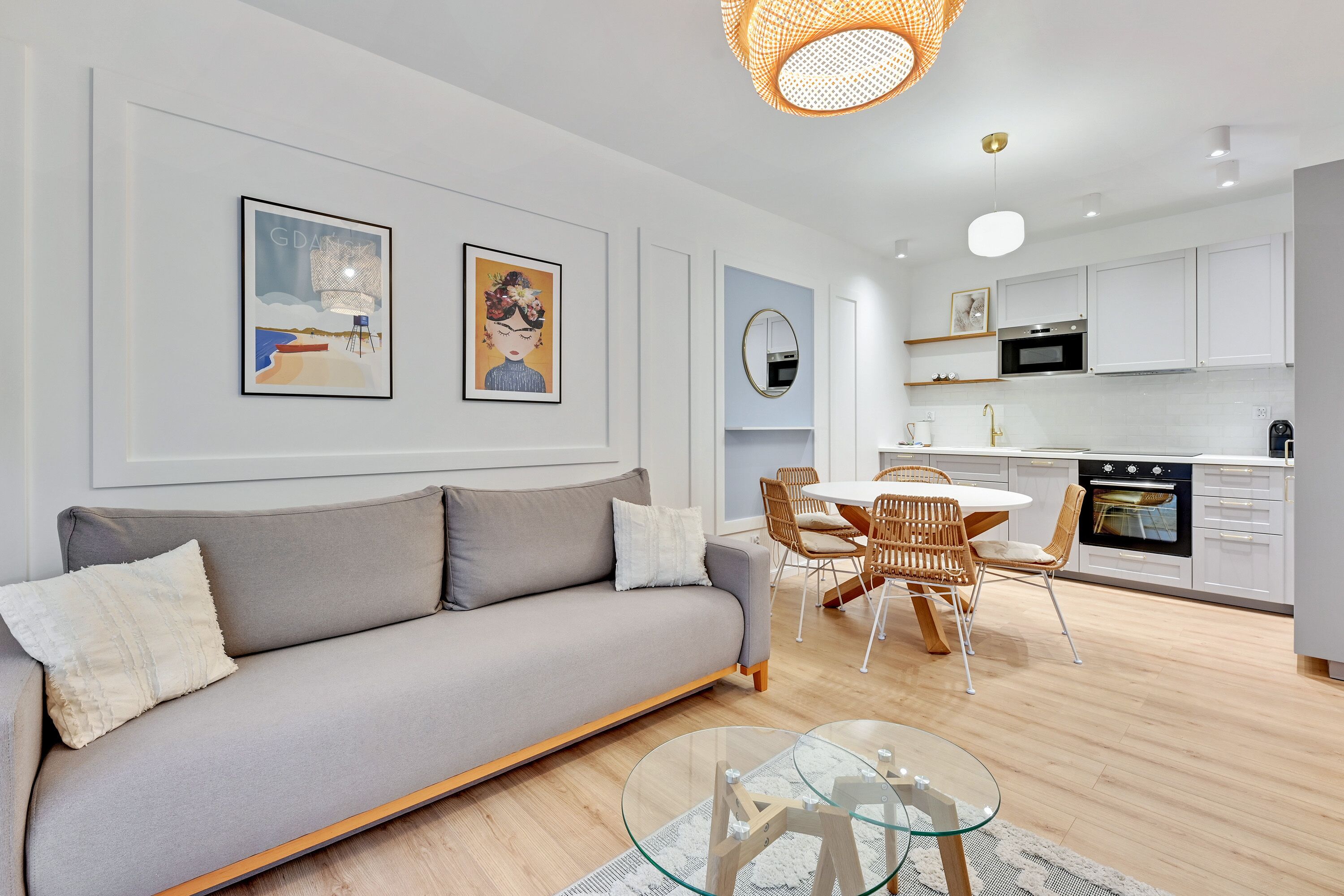 A modern living room with a grey sofa, a glass coffee table, and a dining area with wicker chairs. There is a kitchen in the background with white cabinets and a black oven.