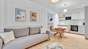 A modern living room with a grey sofa, a glass coffee table, and a dining area with wicker chairs. There is a kitchen in the background with white cabinets and a black oven.