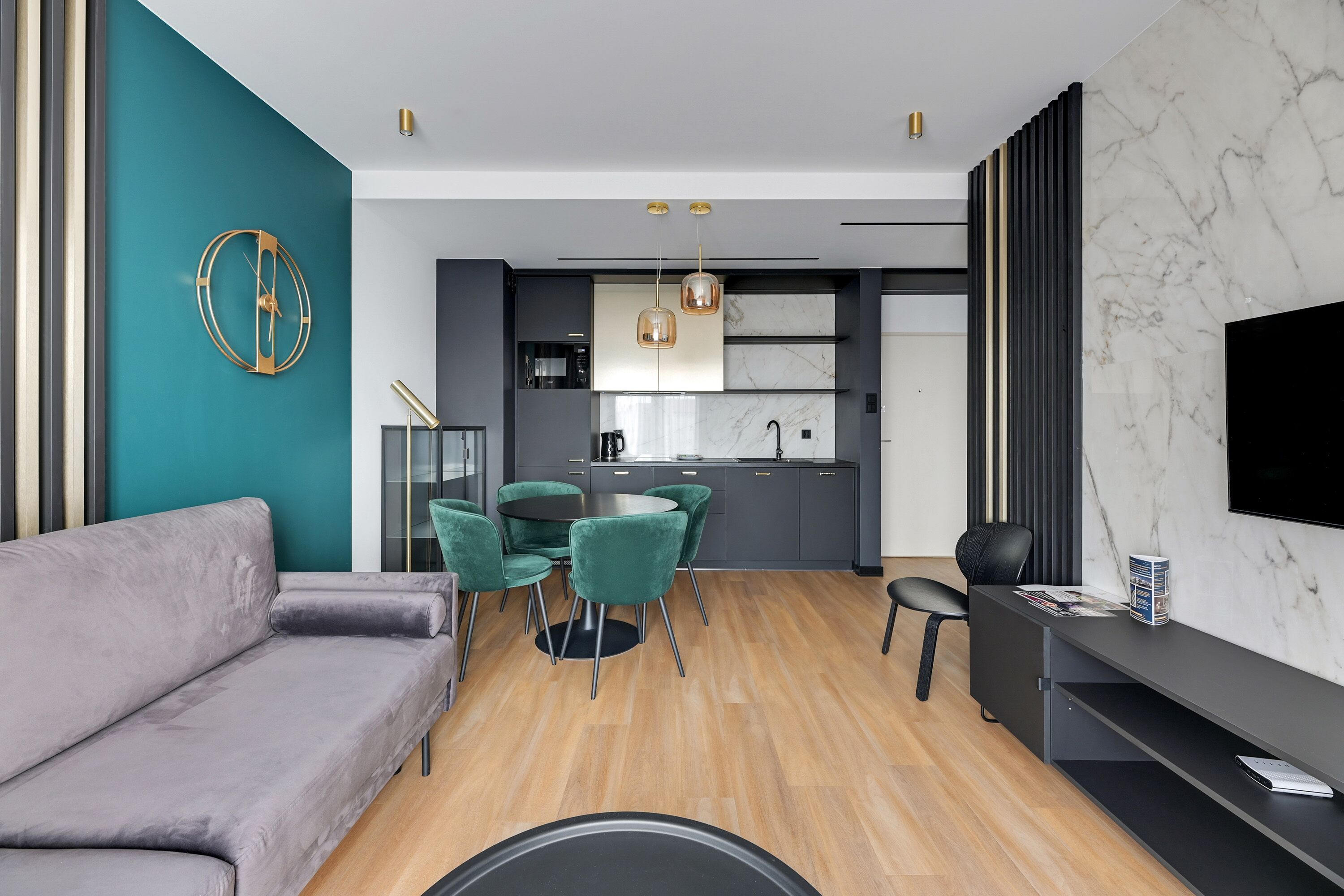 A modern living room with a grey sofa, a dining area with green chairs, and a kitchen with grey cabinets and a marble backsplash.