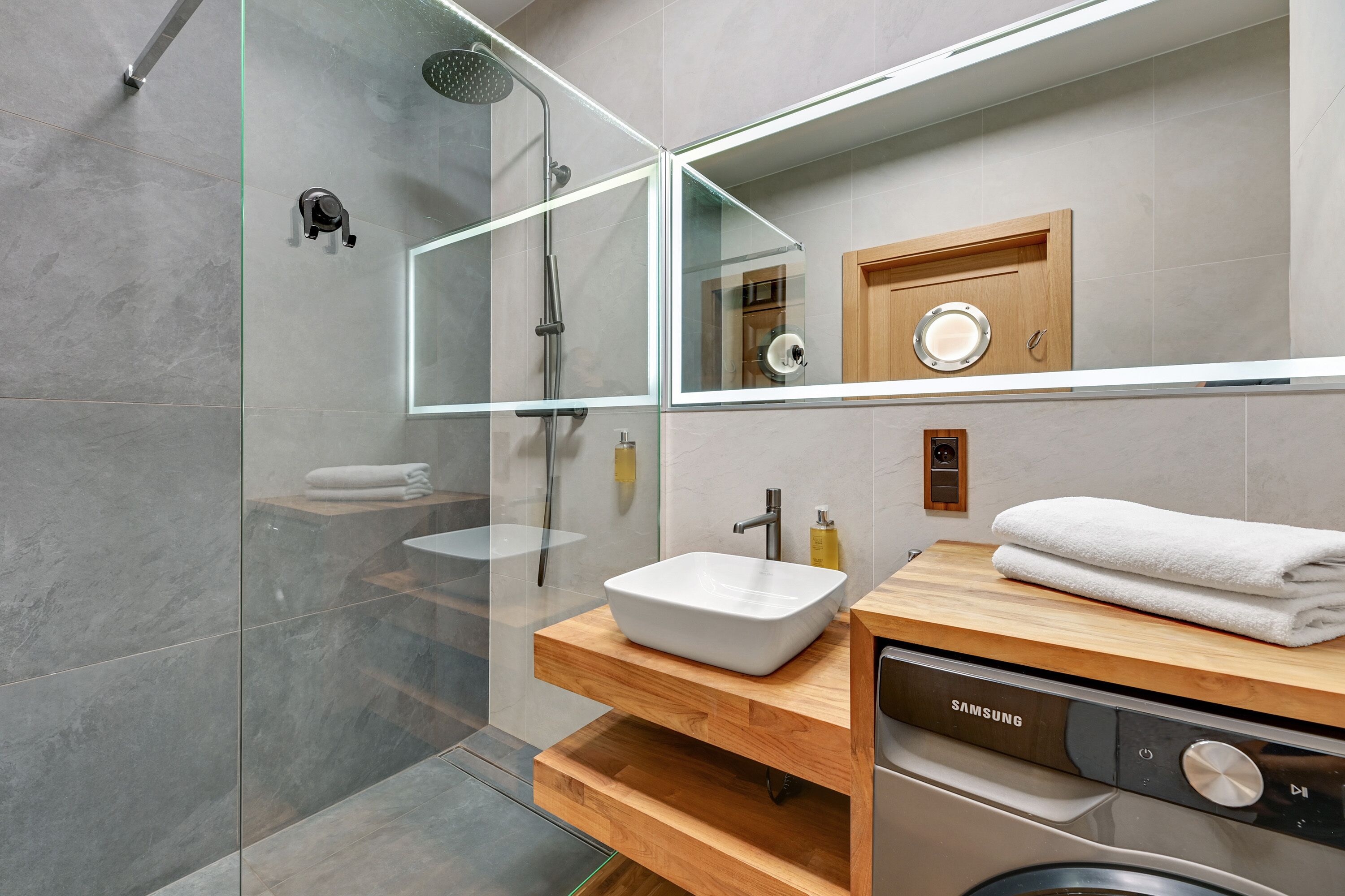 A modern bathroom with a glass-enclosed shower, a wooden vanity with a white sink, and a Samsung washing machine.