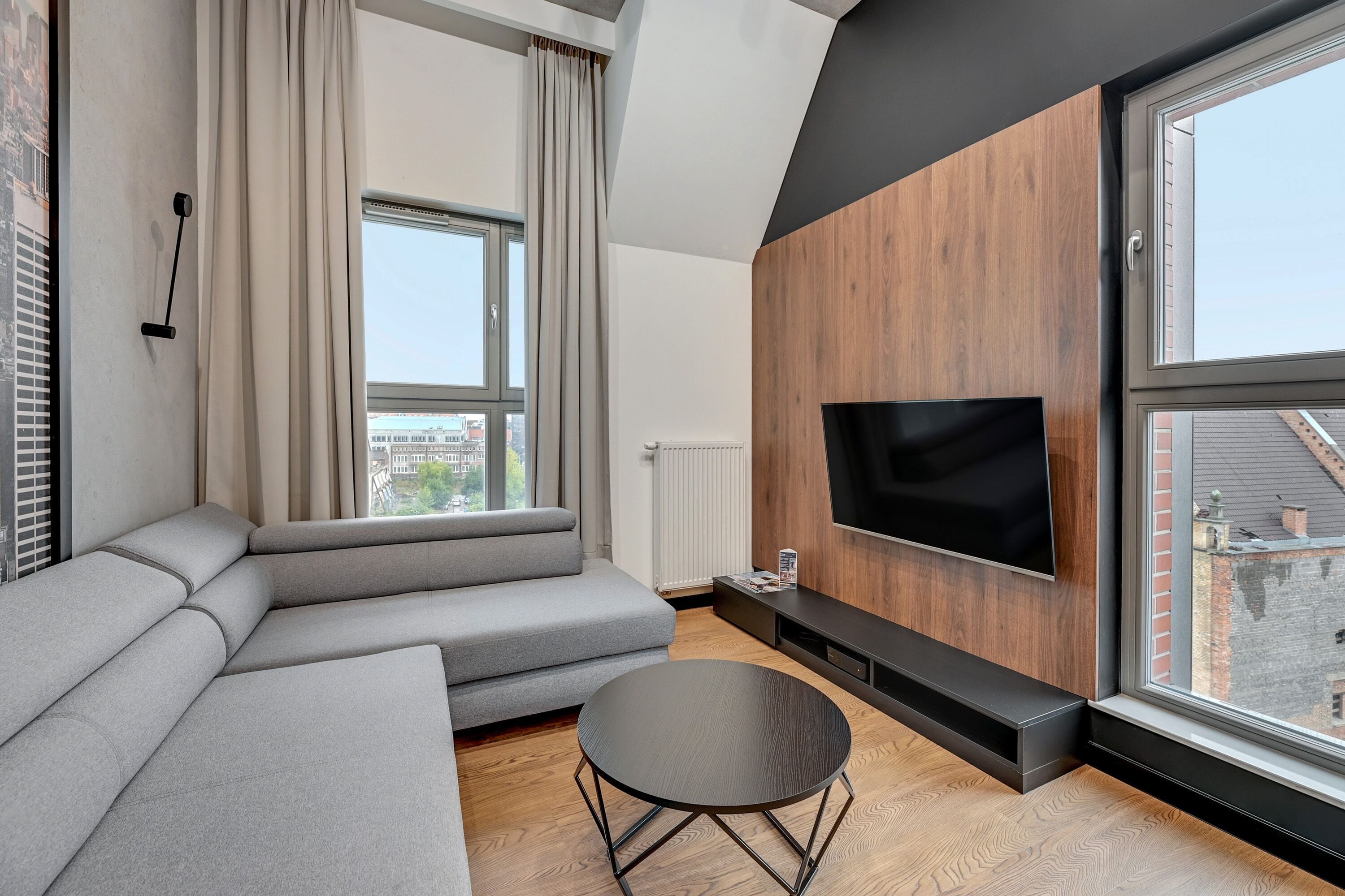 A modern living room with a grey sectional sofa, a round coffee table, a flat-screen TV mounted on a wooden wall, and large windows offering a city view.