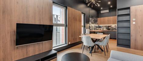 A modern living room with a flat-screen TV, a dining area with a wooden table and chairs, and a kitchen with a marble backsplash.