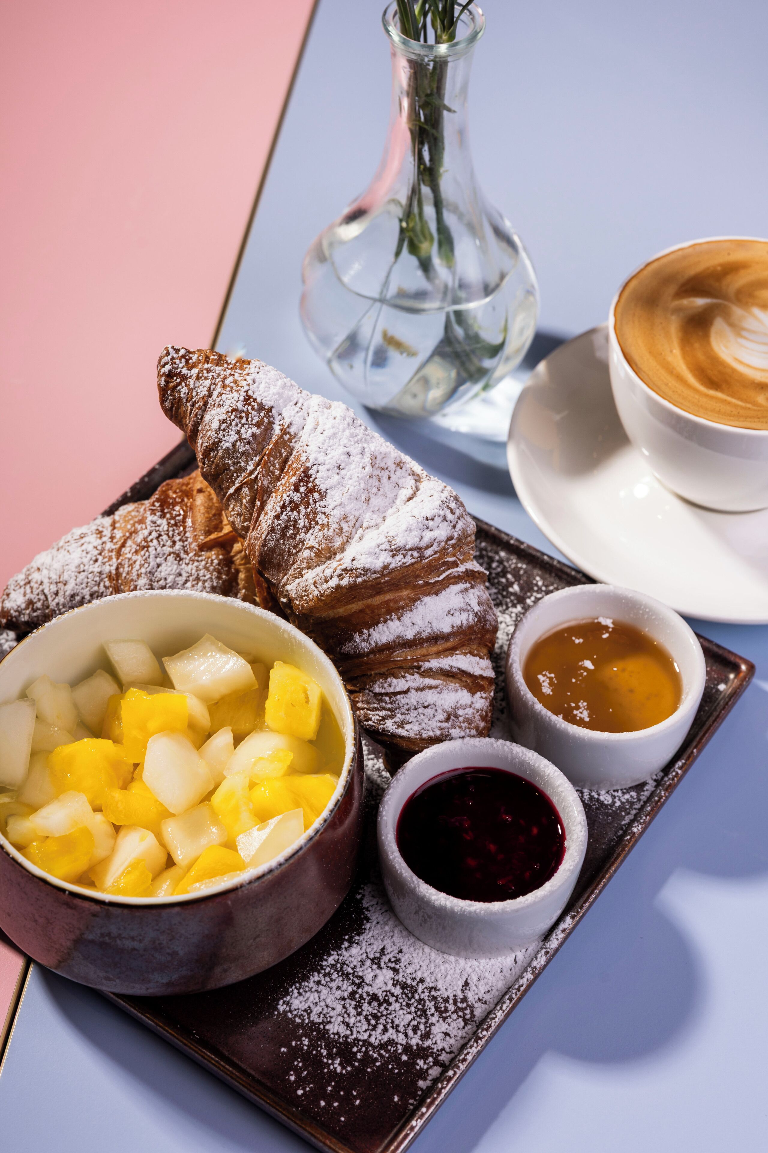 En bakke med en croissant, en skål med frugt samt to små skåle med marmelade og sirup.