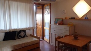 A cozy room with a bed, a table, and a chair. There's a window with curtains, a ceiling fan, and a wall-mounted light.