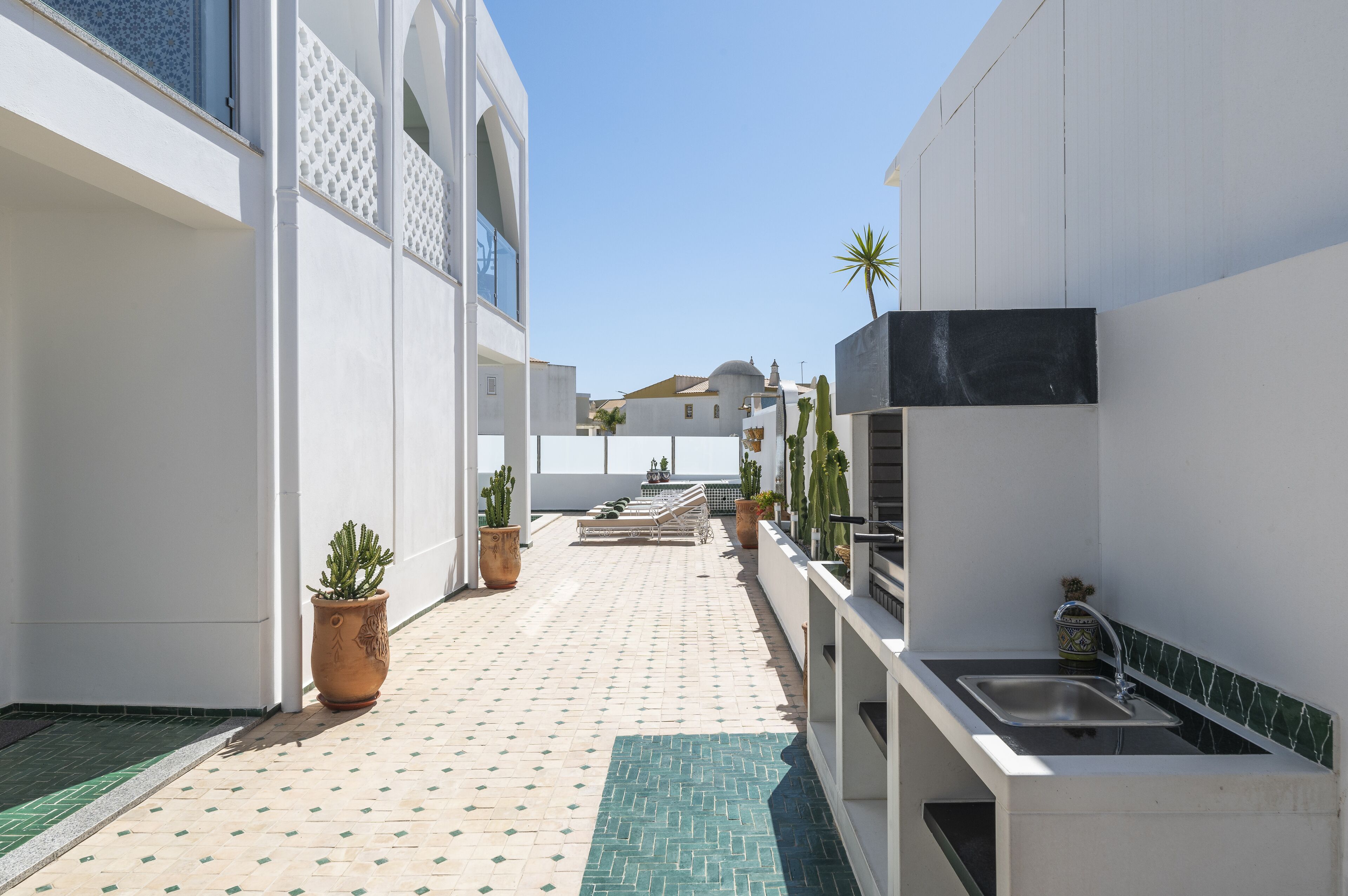 A narrow, tiled corridor with a sink and potted plants, leading to a white building with a tiled roof.