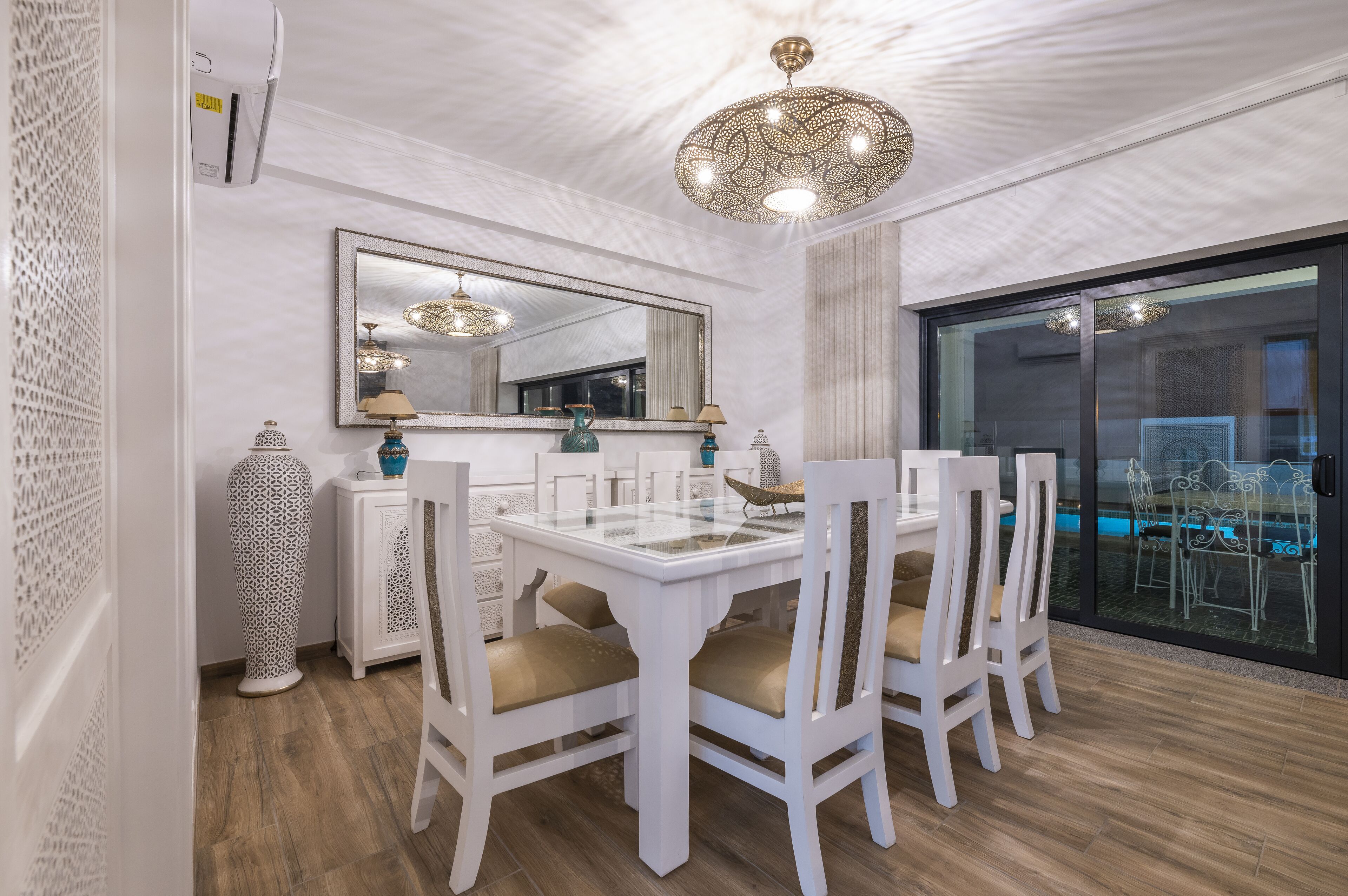 Un coin repas avec une table et des chaises blanches, un grand miroir, des vases décoratifs et un lustre moderne.