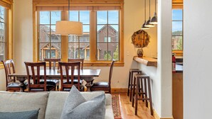 Un coin repas avec une table et des chaises en bois, un lustre et une grande fenêtre donnant sur des maisons.