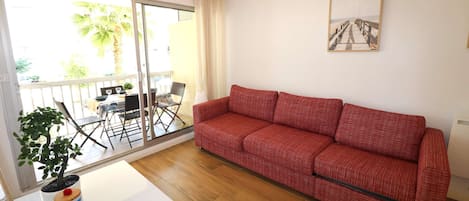 Un sofà vermell, una taula de menjador, cadires, una planta en test i una porta corredissa de vidre que dona a una zona exterior amb una palmera.