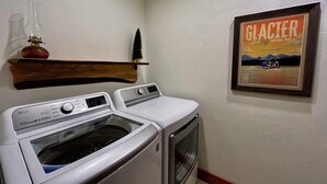 A laundry area with a front-loading washer and dryer, a wooden shelf with a decorative item, and a framed poster of Glacier National Park.