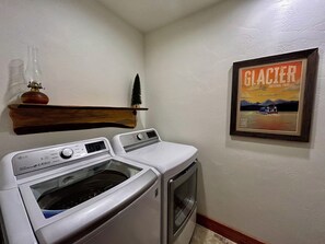 Een wasruimte met een voorlader wasmachine en droger, een houten plank met een decoratief object, en een lijst met een poster van Glacier National Park.