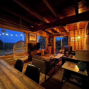 A cozy cabin interior with a wooden dining table, chairs, a hanging light fixture, and a television. - Serene Glacier Cabin With River and Mountain Views.  (Columbia Falls)