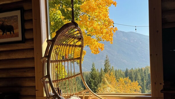 A log cabin interior with a hanging chair by a window overlooking a forest and mountains.