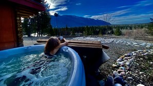 A hot tub with a person relaxing, surrounded by a wooden deck and a view of mountains.