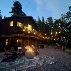 A two-story wooden house with a stone patio, outdoor seating, and a fire pit. - Serene Glacier Cabin With River and Mountain Views.  (Columbia Falls)